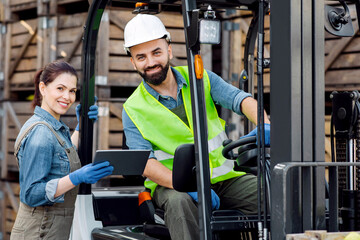 Warehouse workers, industry management with modern technology. Smiling millennial woman with tablet speaks with bearded man in helmet in forklift truck check order, on many wooden boxes background