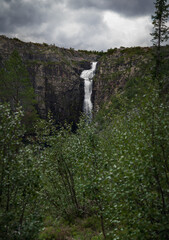 Njupeskär Waterfall flows dramatically down a rugged cliff in Fulufjället National Park, surrounded by dense foliage and under a moody, overcast sky. In Dalarna Sweden
