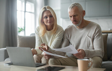 A senior couple sits on a sofa in a bright room as they go through financial papers together. They...