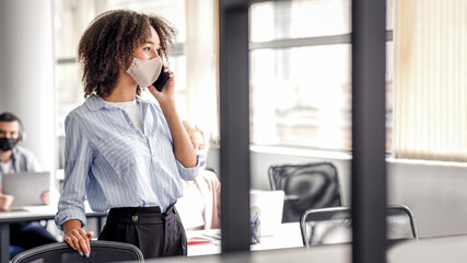 Manager speaks on phone with client. Attractive african american millennial lady in protective mask talk on smartphone while discussing business issues in office interior during covid-19 epidemic