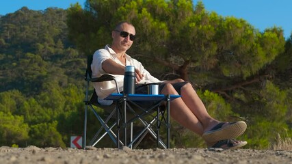 Savoring a warm beverage from a thermos. Tourist is relaxing in a folding chair by a roadside, enjoying a hot drink from a thermos. He is taking a break from driving and enjoying the scenery