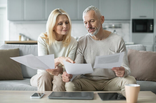 A senior couple sits closely on a sofa, reviewing various documents while discussing important matters. Morning sunlight fills the cozy living room, enhancing their focused interaction.