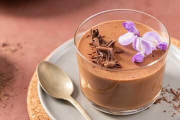 A delicious individual chocolate mousse in a glass, topped with chocolate shavings and purple flowers, presented on a plate with a spoon.