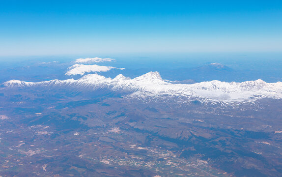 Aerial view of stunning mountain range with snowy peaks under blue sky taken during flight. Below mountains, vast landscape of rolling hills, scattered settlements stretches out blending into horizon
