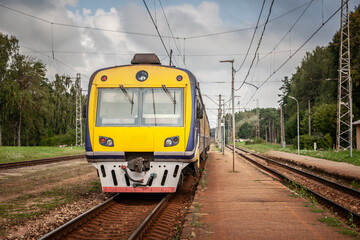 Obraz premium Yellow-blue Russian made passenger train at a rural station platform in Saulkrasti, Latvia; overhead wires, twin tracks and forest edge illustrate typical Eastern-European commuter rail.