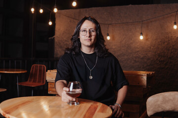 young latin man with long hair holding a glass of beer in a bar while looking at the camera