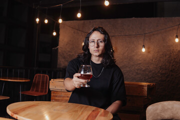 young latin man with long hair holding a glass of beer in a bar while looking at the camera