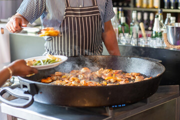 Chef cooking shrimp and vegetables in a large pan at an outdoor event in the afternoon sunlight