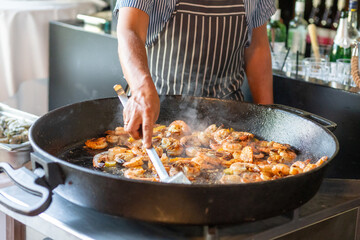 Chef prepares a seafood dish with shrimp in a large pan at a bustling kitchen during a lively evening event