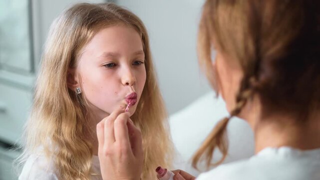 Close-up of girl with blonde hair pouting her lips while mother applies pink lip gloss. Warm beauty care moment captures joy, fun and connection in family lifestyle at home.