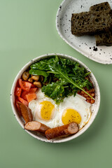 Vibrant breakfast bowl combines perfectly cooked eggs, fresh greens, and sliced sausage, served alongside a plate of rustic bread. A wholesome start to the day