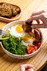 A colorful breakfast showcases perfectly cooked eggs beside fresh greens and ripe tomatoes. Crusty homemade bread complements the dish as hands reach in to enjoy a comforting morning feast