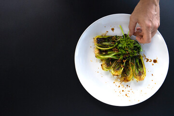 Hand adjusts a plate of bok choy garnished with seasoning, perfect for Asian cooking.