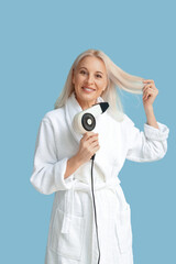 Fototapeta premium Mature woman in bathrobe with hair dryer on color background