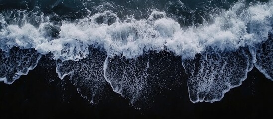 Drone aerial view of the sea or ocean waves splashing on a dark navy blue or gray sand beach with beautiful transparent clear water