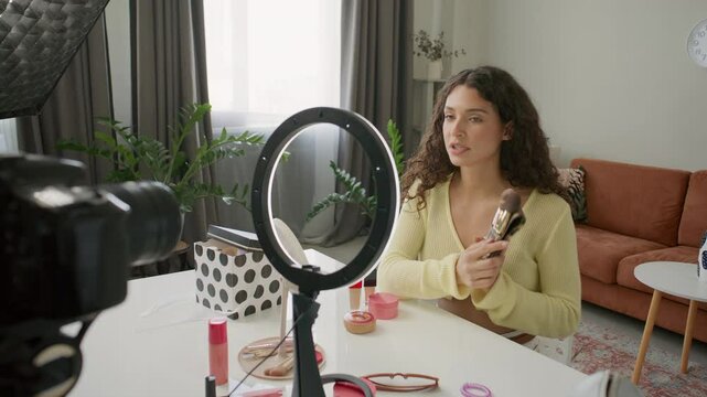 Female beauty blogger sitting at desk in home studio, presenting brush set while recording makeup tutorial or review with professional digital camera and ring light