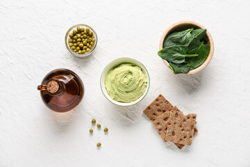 Bowl with tasty peas hummus, spinach, jug of oil and crackers on light background