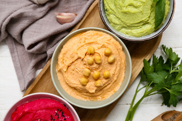 Bowls of different tasty hummus with garlic, parsley, beet and crackers on white wooden background