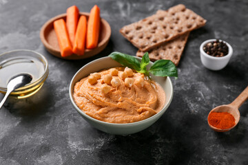 Bowl of tasty hummus with carrot, chickpeas and crackers on black grunge background