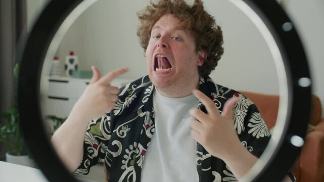 Young male speech coach pointing at mouth and explaining pronunciation and articulation techniques, recording video tutorial with ring light setup at home