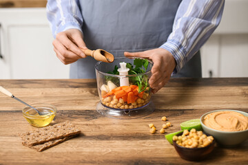 Woman adding spices into blender with chickpeas, carrot and parsley for hummus at table in kitchen