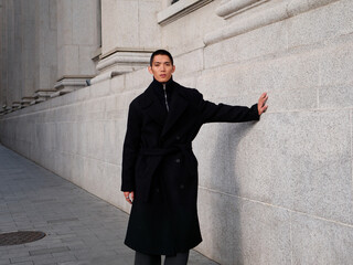 Portrait of handsome Chinese young man wearing black overcoat posing in the street, young guy with black short hair all in black with urban background. Male fashion, cool Asian young man lifestyle.