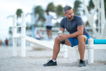Sport fitness man resting on a bench outdoors. Mature fitness sport model sitting on a bench after a workout. Sports man taking a break after fitness. Sport guy resting after fitness. Sport for adult.