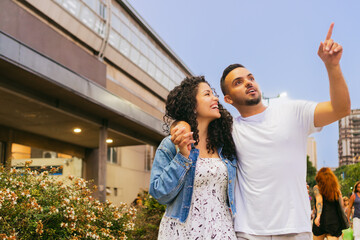 Young Latin man, hugging his partner, points to the distance, sightseeing, surrounded by flowers at street, with copy space
