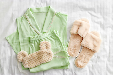 Top view of folded light green woman pajama, fluffy fur sleep eye mask, soft home slippers on white sheet bed. Minimal flat lay cozy comfort pyjamas. Aesthetic lifestyle photo of stylish sleepwear