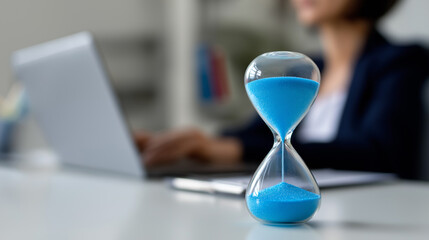 Blue sand hourglass on a desk with a businesswoman working on a laptop in the background.
