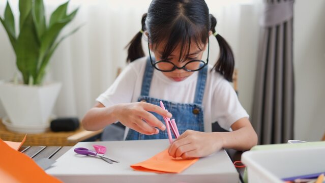 Asian girl focused on making a DIY craft project at home using a compass and colorful paper, promoting creativity, fine motor skills, and fun learning in a homeschool setting.