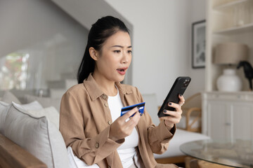 Asian woman stare at cellphone screen with shocked expression while holding debit card, experiencing technical issues, declined order due to insufficient funds, payment failure or security concerns