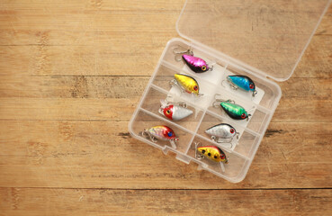 Fishing lures are in a plastic box on a wooden floor with space for copying.