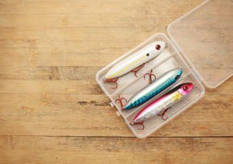 Fishing lures are in a plastic box on a wooden floor with space for copying.