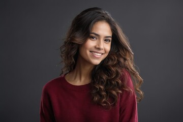 Confident Hispanic woman with long hair, smiling softly in a maroon sweater against a plain gray setting.

