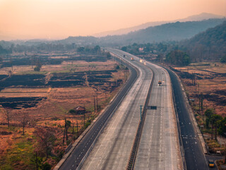 Aerial view of a scenic highway, part of the Mumbai-Goa Highway, stretching through rural fields and hills at sunset, with vehicles travelling along the road under a warm, orange sky.