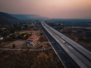 Aerial view of a scenic highway, part of the Mumbai-Goa Highway, stretching through rural fields and hills at sunset, with vehicles travelling along the road under a warm, orange sky.