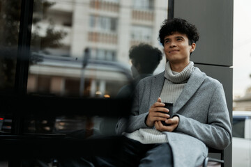 Young man sitting on bench in front of building with hands in pockets and cup of coffee in hand