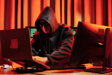 A man in a dark hoodie uses a laptop at night, symbolizing cybercrime, hacking, identity theft, and...