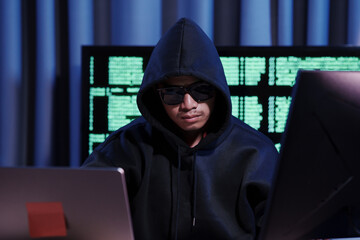 A man in a dark hoodie uses a laptop at night, symbolizing cybercrime, hacking, identity theft, and...