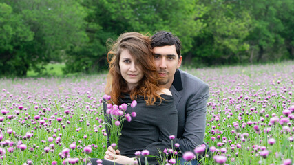 Loving couple sitting in a field of purple wildflowers. Woman holds flowers while man embraces her...