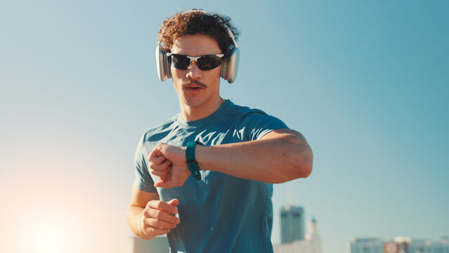 Man, fitness and checking watch with headphones in city for workout app or tracking performance. Active, male person or runner with wristwatch for music, calories or running exercise on space in town