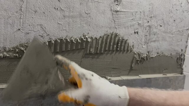 A professional construction worker is applying tile adhesive to a bathroom wall. This is the process of spreading the mortar with a notched trowel and preparing the surface before laying the tiles.