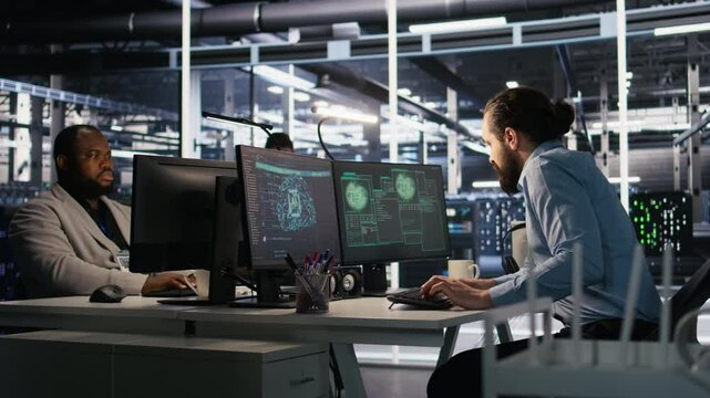 Engineer in server room responding to sudden ransomware detection on monitor, requesting help from manager to protect critical data. Worker reviewing suspicious activity logs, informing team leader