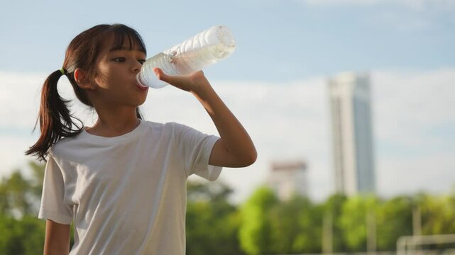 Asian young girl drinking water after outdoor activity in the sunlight, promoting hydration, child wellness, and healthy habits through physical care and active lifestyle.