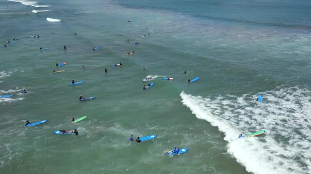 Aerial view of surf lessons with surfers paddling and riding small waves near the shore in Bali, Indonesia.
