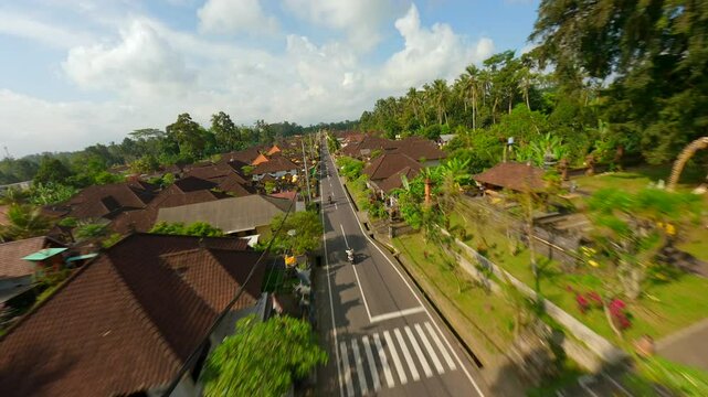 Cinematic FPV drone shot of motorbike riding through decorated street in traditional Balinese village surrounded by lush tropical greenery in Bali, Indonesia. 