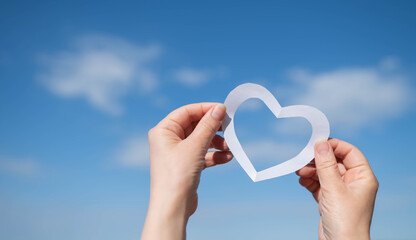 A woman's hand holds a white paper heart against a clear blue sky and lake, symbolizing love and...