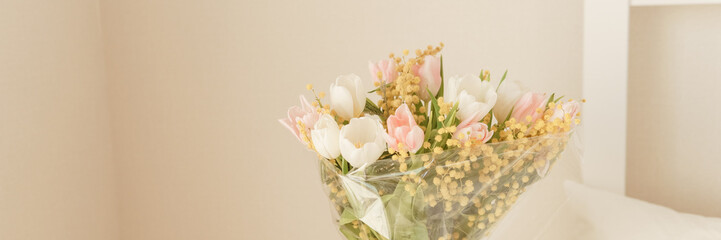 Bouquet of pink and white tulips with mimosa in a glass vase in a bedroom. Spring flower arrangement for interior decoration.