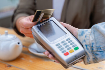 Close-up view of a person making a contactless payment using a credit card on a POS terminal. The scene takes place in a cafe setting with a teapot in the background, showing modern transaction ease 